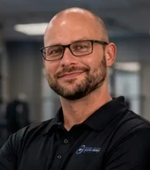 Bald man with glasses and beard smiling in black polo shirt